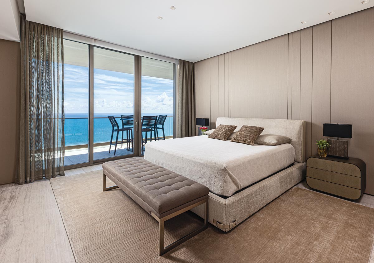 Underscoring the exceptional water vistas with a breezy outdoor balcony, the primary bedroom maintains the home’s subdued design with a platform bed and headboard from Fendi Casa. A tufted Artefacto bench amplifies the room’s elegant simplicity, and gossamer draperies frame the view.